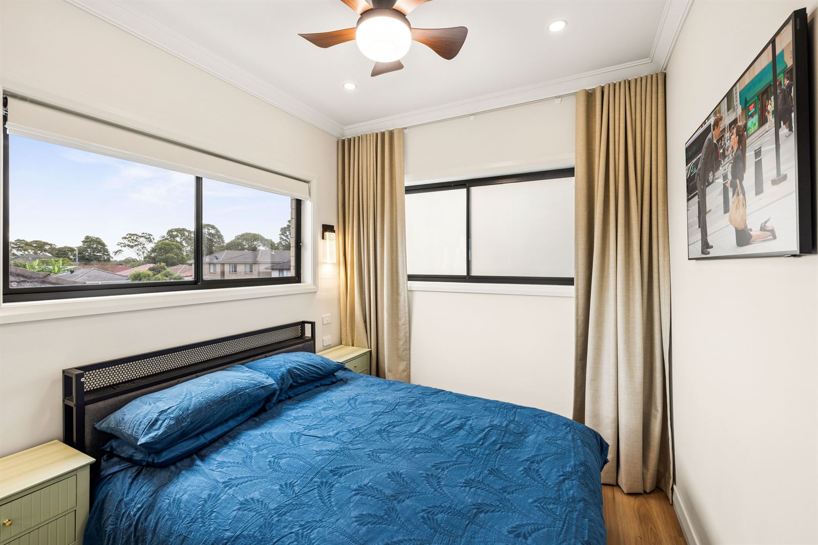 Bedroom with a queen bed, ceiling fan and a wide window over the neighbouring rooftops.